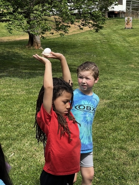 Two children stand on grass outdoors, one raising a small object above their head while another stands beside them watching. Trees and open space are visible in the background.
