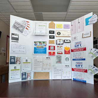 A tri‑fold display board set up on a table, covered with various gift certificates, business cards, flyers, and promotional materials from local businesses. Items include recognizable logos such as McDonald’s, Starbucks, and MFA Oil, along with envelopes and printed certificates arranged neatly across the board. The display appears to showcase raffle or giveaway prizes for a community or school event.