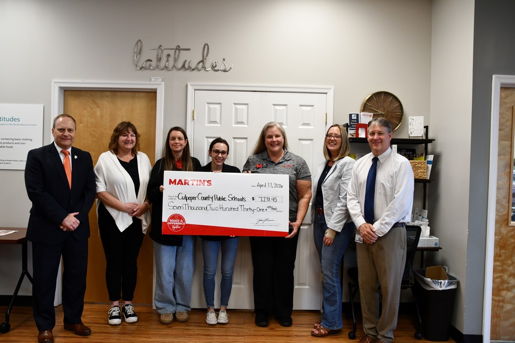 A group of seven adults stand indoors in front of double white doors beneath a wall sign reading “latitudes.” Three women in the center hold a large ceremonial check from MARTIN’S made out to Culpeper County Public Schools for $7,231.45, dated April 17, 2020. The group is smiling and dressed in business-casual attire, with one man in a suit on the far left and another on the far right. A wooden door, wall clock, shelving unit, and office items are visible in the background, suggesting a community resource or office setting.