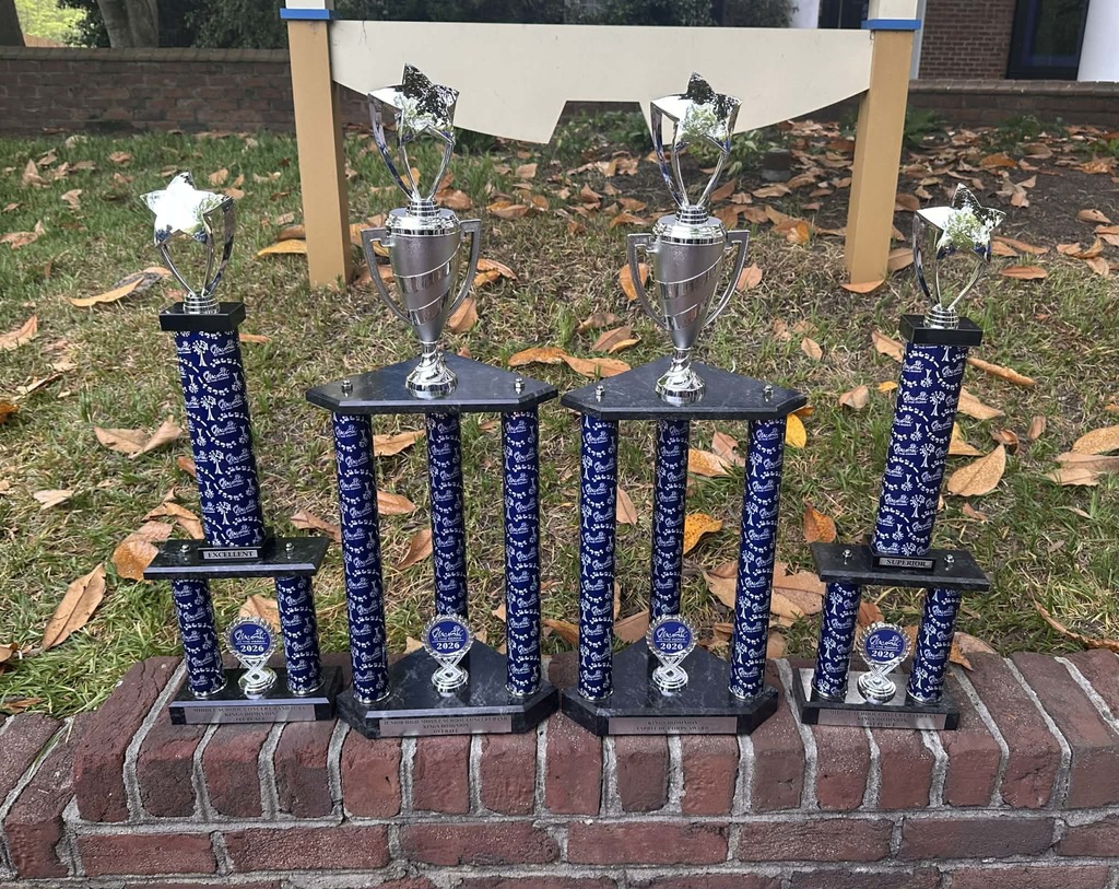 A row of four trophies displayed outdoors on a brick ledge, featuring two tall first-place trophies, a larger grand champion-style trophy, and two additional awards with star toppers, recognizing a school band’s competition achievements.