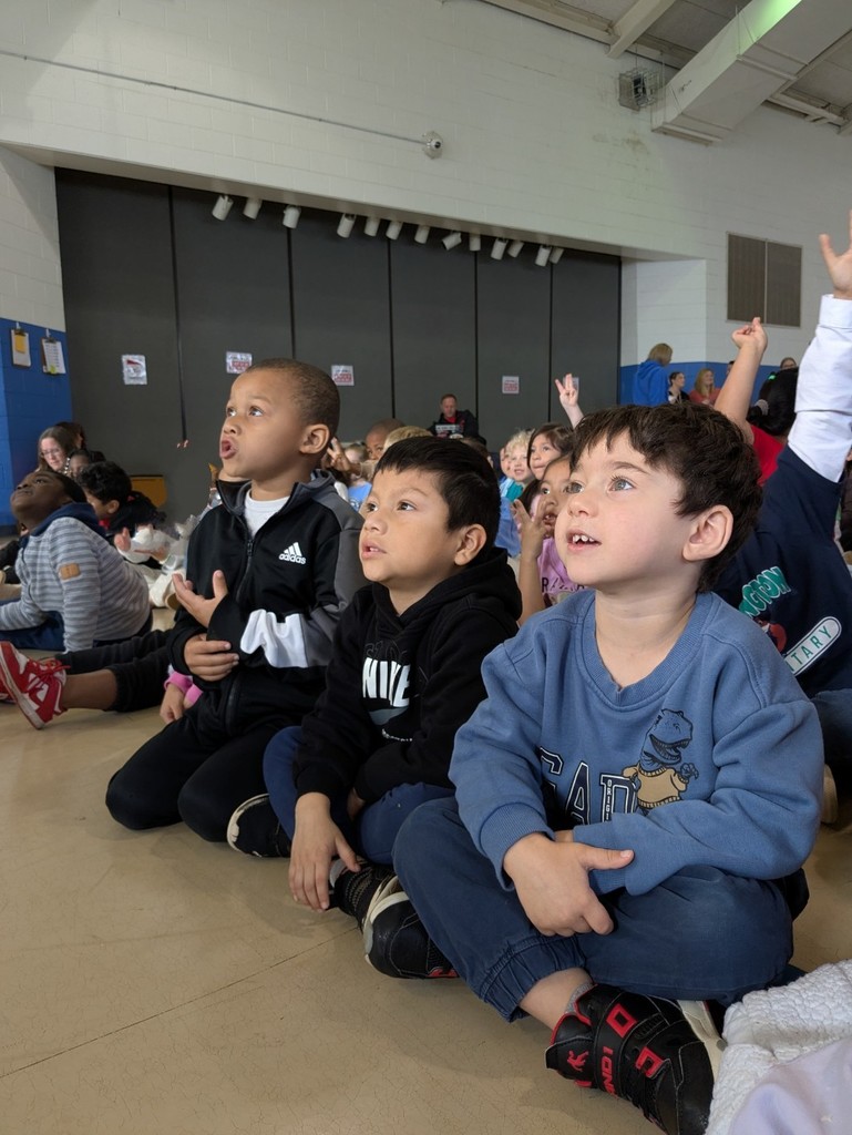 A group of young students sit closely together on a gym floor, looking upward with curiosity and excitement during a live science presentation.