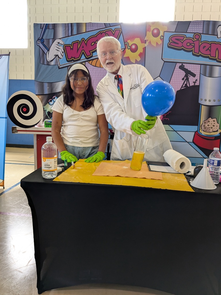 A science presenter in a lab coat holds a bottle with a balloon inflating on top while a student wearing safety glasses stands beside him at a demonstration table.
