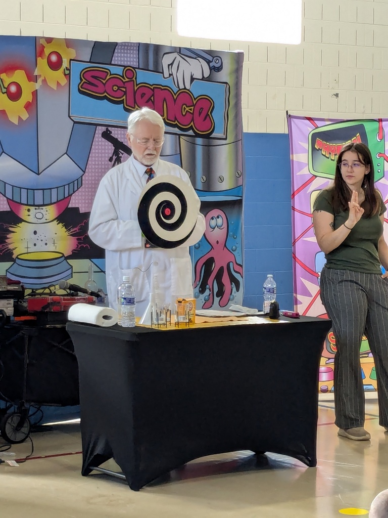 A man in a lab coat stands behind a table demonstrating a science experiment with a spiral target prop while a woman stands nearby assisting in a school gym.