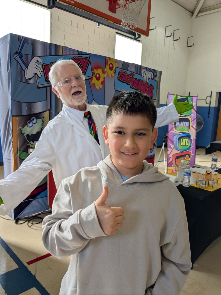 A smiling elementary-age boy in a light hoodie gives a thumbs-up in a school gym while a white-haired man in a lab coat and safety glasses gestures behind him in front of a colorful “Wacky Science” backdrop.
