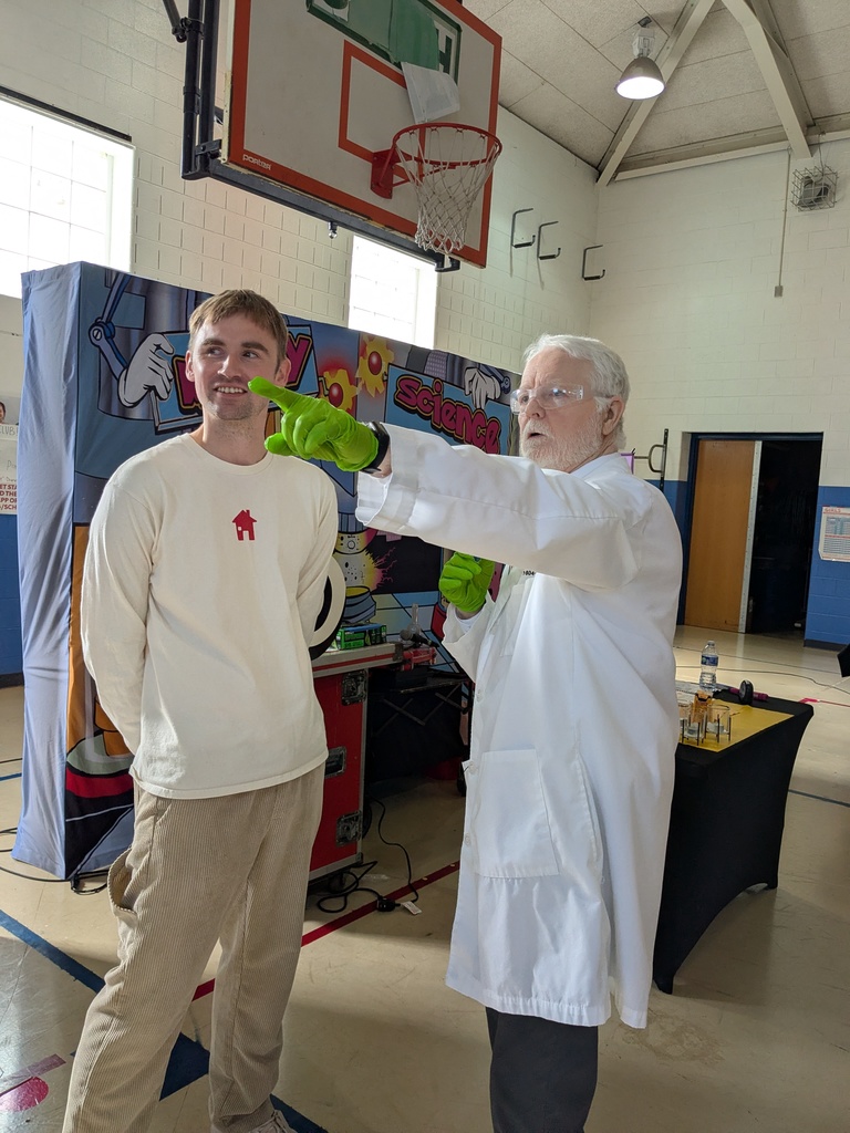 An older man in a lab coat and bright green gloves points across a school gym while speaking to a younger adult standing beside him near a science demonstration setup.