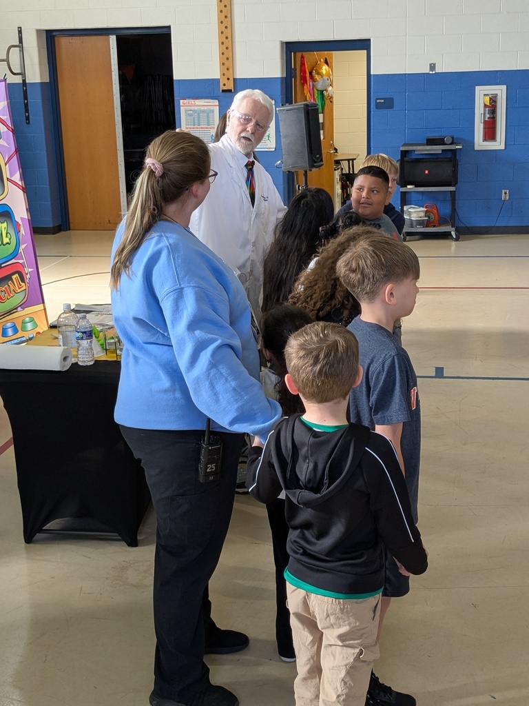 A man in a lab coat interacts with a small group of elementary students gathered around him in a gym during a hands-on science activity.