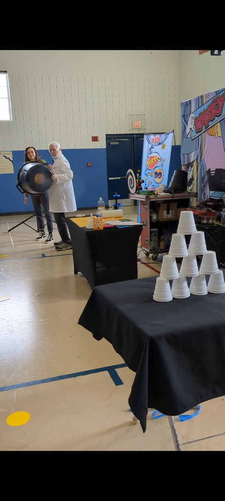 A science presenter and assistant stand near demonstration equipment in a gym, including a large gong-like instrument and stacked cups on a table in the foreground.