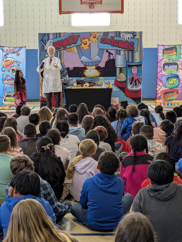 A wide view of a school gym shows a science presenter performing on stage in front of a large seated audience of elementary students and a colorful “Wacky Science” backdrop.