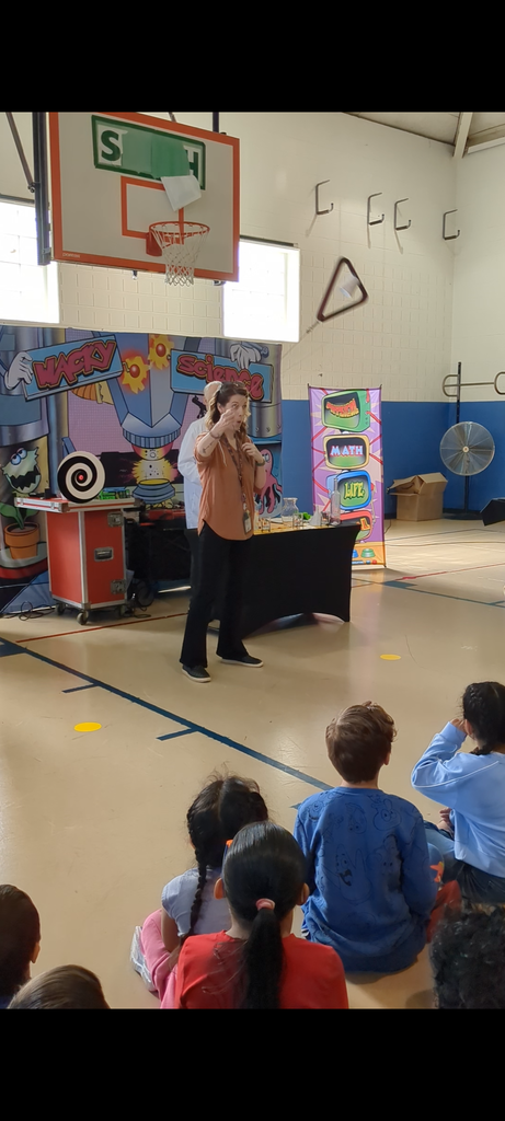 A triangular metal instrument is midair as a woman gestures toward the audience during an interactive science demonstration in a school gym.