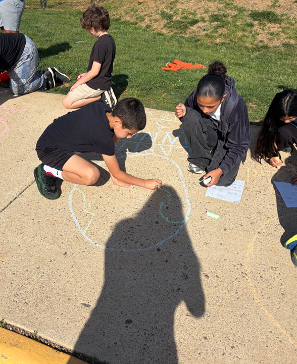 Students kneel on the sidewalk drawing and labeling planets with chalk, adding details and fun facts as part of a hands-on solar system lesson.