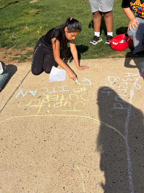 Children spread out along a curb outside the school, each drawing a planet in chalk and writing a fact, continuing a classroom activity about the solar system.
