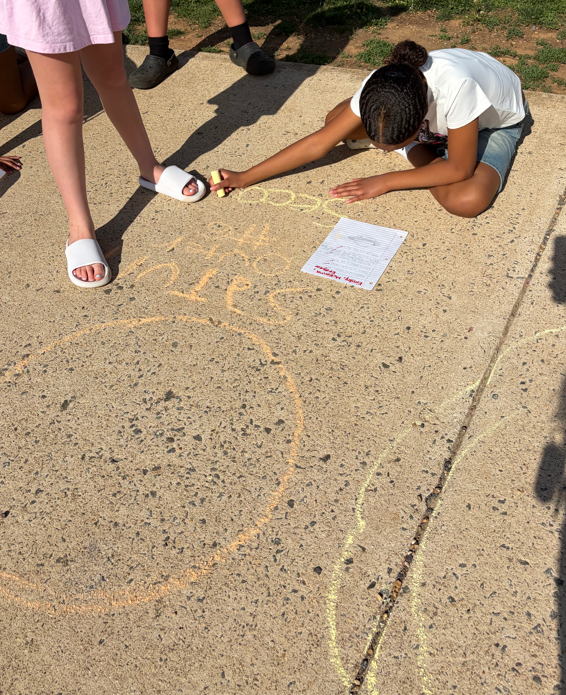 Elementary students work together on a sidewalk, using chalk to draw a large planet and write facts after researching the solar system in class.