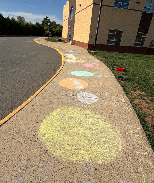 Sidewalk outside a school building covered in colorful chalk drawings of the planets arranged in order like the solar system, created by students during an outdoor science activity.