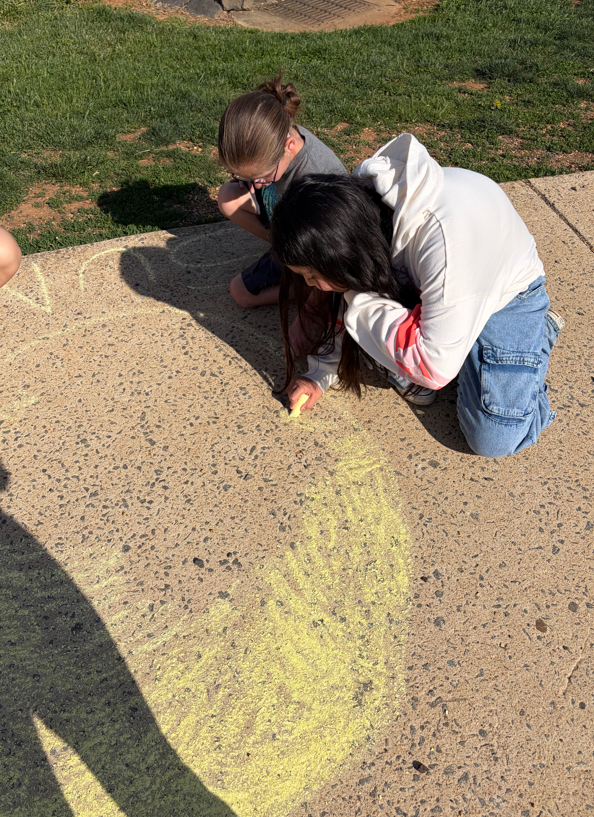 Groups of students create chalk drawings of planets inside large circles on the sidewalk, with research papers nearby, combining science learning with outdoor art.