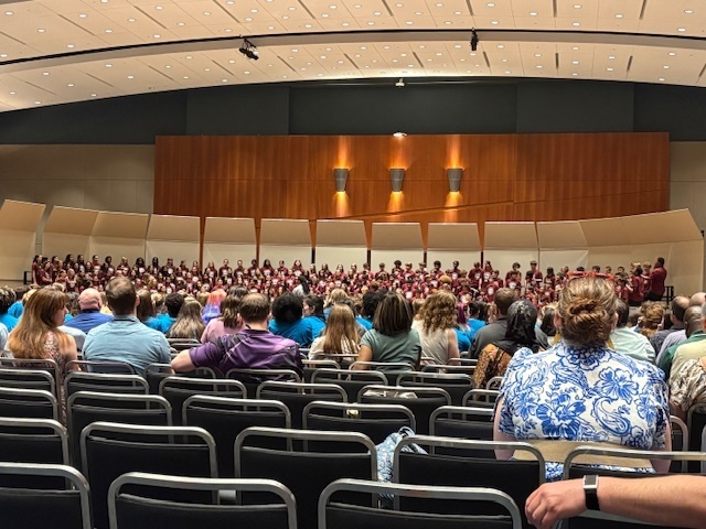 A large middle school choir performs on stage in a concert hall, with rows of audience members seated and watching the performance.