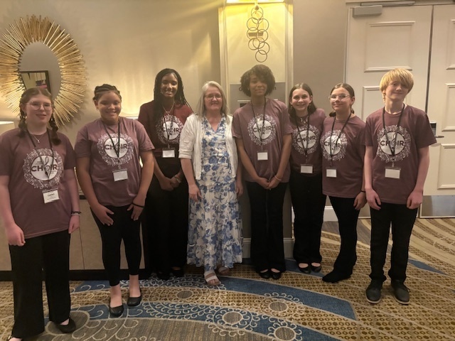 A group of middle school students wearing matching choir shirts stand with an adult in a hotel or conference setting, smiling and posing together.