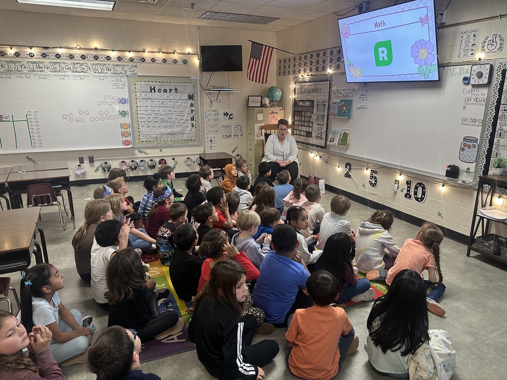A classroom of young students sitting together on a colorful rug, facing a teacher who is seated at the front. The teacher is leading a lesson while a large screen displays a math activity. The room is decorated with educational posters, string lights, and classroom materials, creating a warm, organized learning environment.