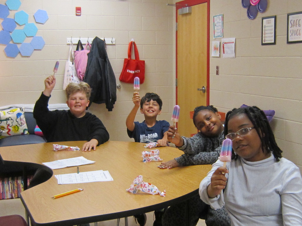 A group of four elementary-aged students sitting around a table in a classroom, smiling and holding up red, white, and blue popsicles; papers, pencils, and wrappers are on the table, with coats and bags hanging on the wall behind them.