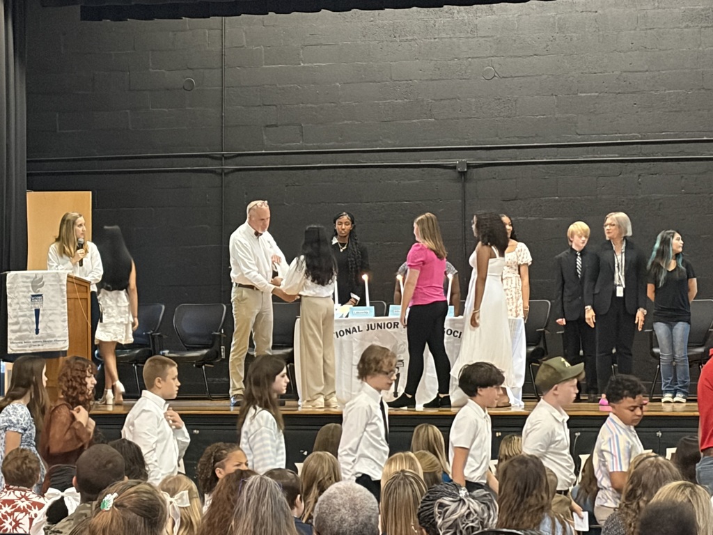 Multiple students line up on stage during a National Junior Honor Society induction ceremony as they take turns receiving recognition, with an audience seated in front.
