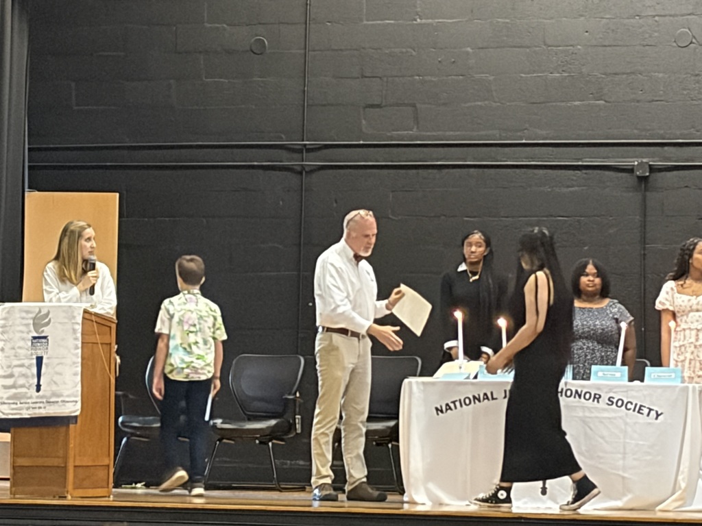Student participates in a National Junior Honor Society ceremony on a stage, receiving recognition near a table with lit candles while a speaker stands at a podium nearby.