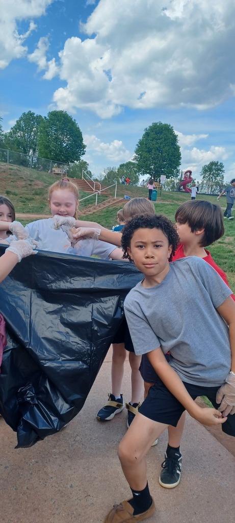 Children wearing gloves collect trash into a large black garbage bag at a park, with a playground and grassy hill in the background under a partly cloudy sky.