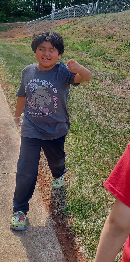 A smiling child walks along a paved path holding a small piece of trash, participating in a park cleanup activity.