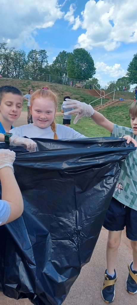 A group of children wearing disposable gloves hold open a large black trash bag while cleaning up litter outdoors on a sunny day.