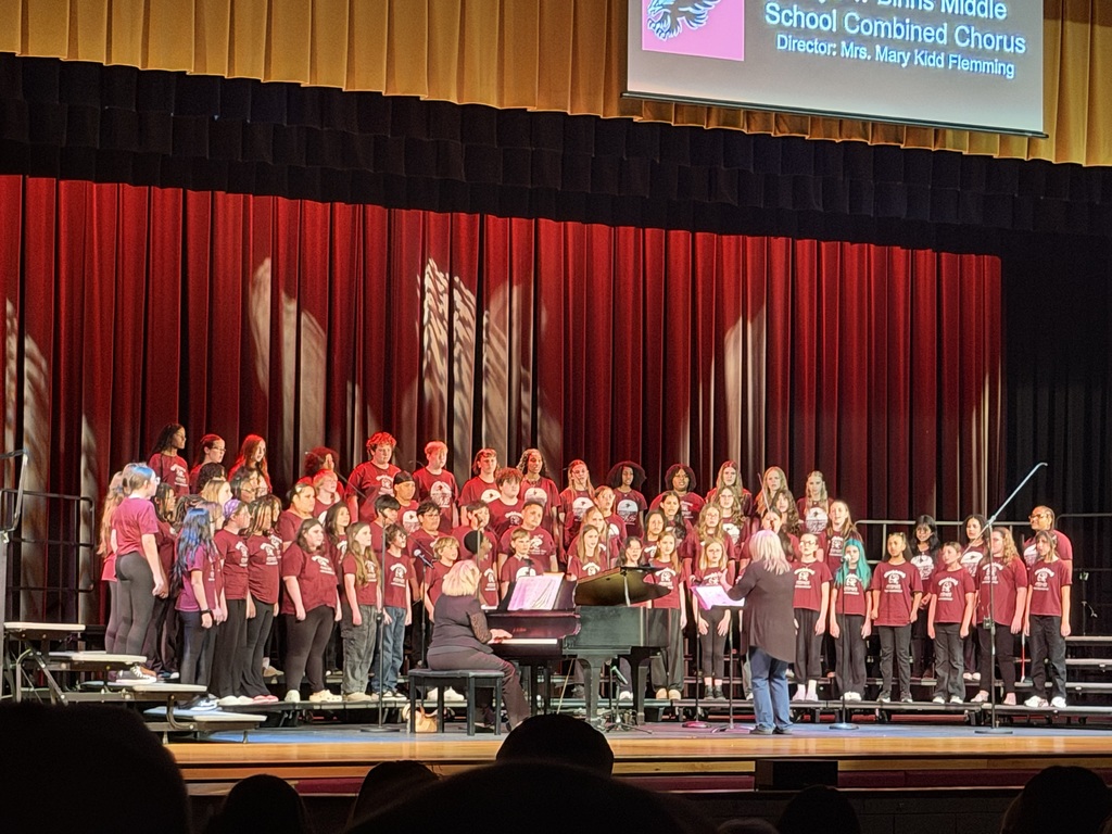 Full middle school combined chorus stands on risers singing on stage while a director leads them and a pianist accompanies.