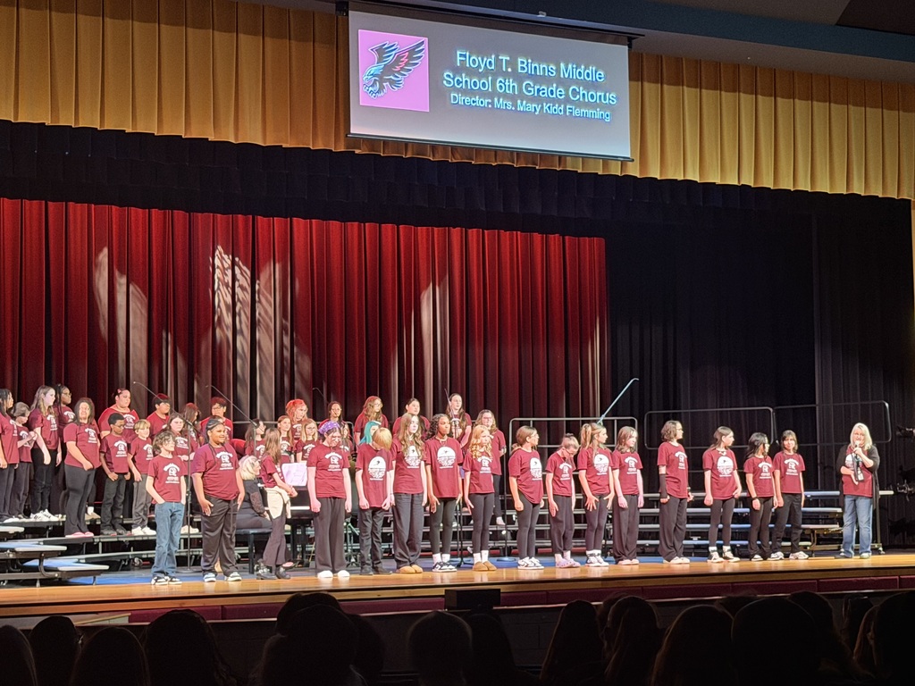 6th grade chorus from Floyd T. Binns Middle School stands in rows on stage as their director speaks into a microphone during a concert.