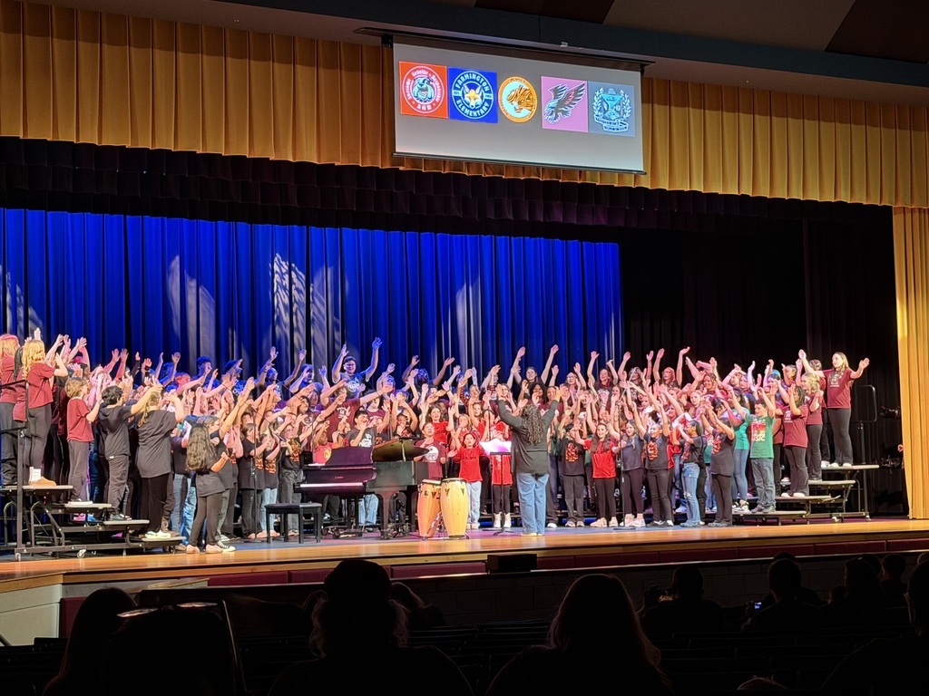 Large group of middle school choir students on stage with their director, raising their arms together during a lively performance under colorful stage lighting.