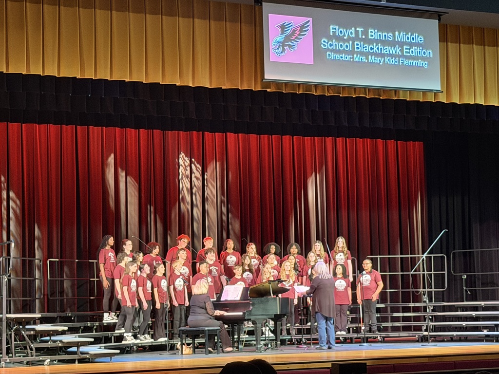 Middle school choir labeled “Floyd T. Binns Middle School Blackhawk Edition” performs on stage with a pianist and director in front of red curtains.