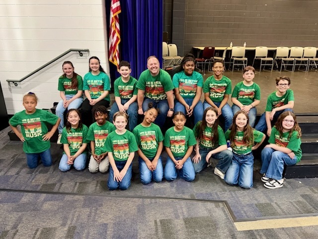 A group of elementary school students and an adult sit and kneel in rows on a stage, all wearing matching green “Life is Better with Music” shirts. They face the camera and smile, posing together in a school auditorium setting with chairs and a flag in the background.