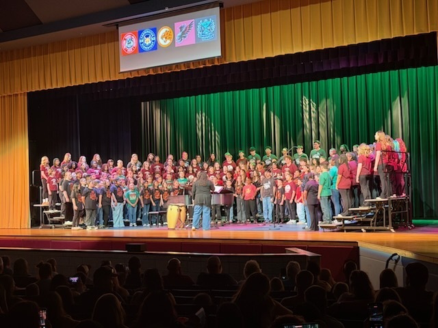 A large group of elementary, middle, and high school students stand on risers in an auditorium, performing together in a chorus concert. A conductor stands in front of them, and a seated audience watches from below. Colorful stage curtains and a projection screen with school logos are visible above the performers.