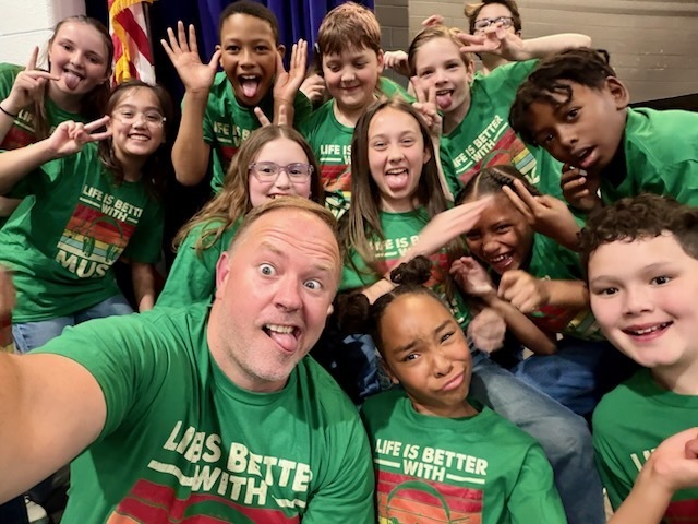 A group of elementary school students and an adult pose closely together for a selfie, all wearing matching green shirts that read “Life is Better with Music.” The group smiles, laughs, and makes playful faces, showing excitement and camaraderie.