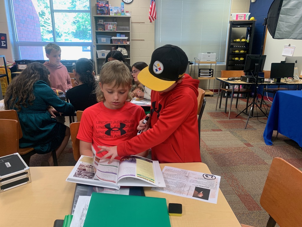 Two students sit side by side at a classroom table, working together to read a textbook and review printed materials. One student points to a page while the other follows along, as additional groups collaborate at tables in the background.
