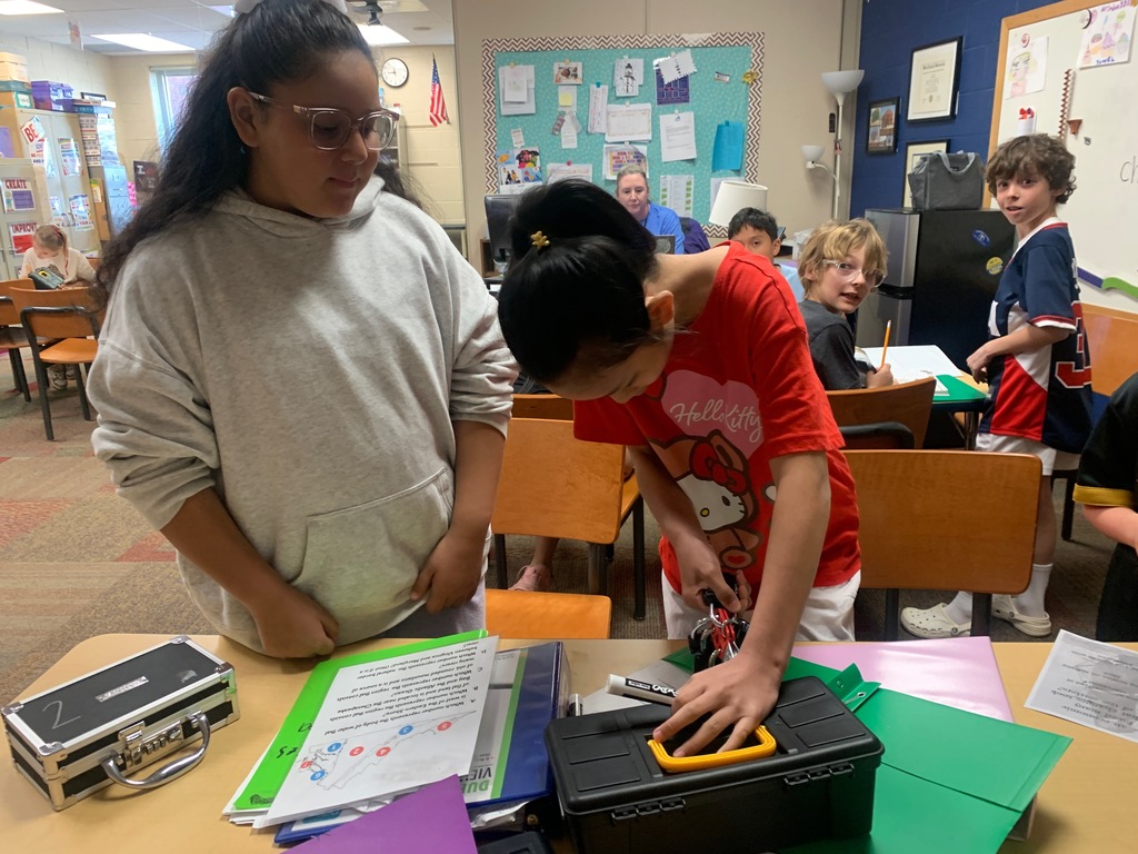 Two students collaborate at a desk, using a key and lock to open a small black box while reviewing written instructions and materials spread across the table. Other classmates sit and watch in the background.