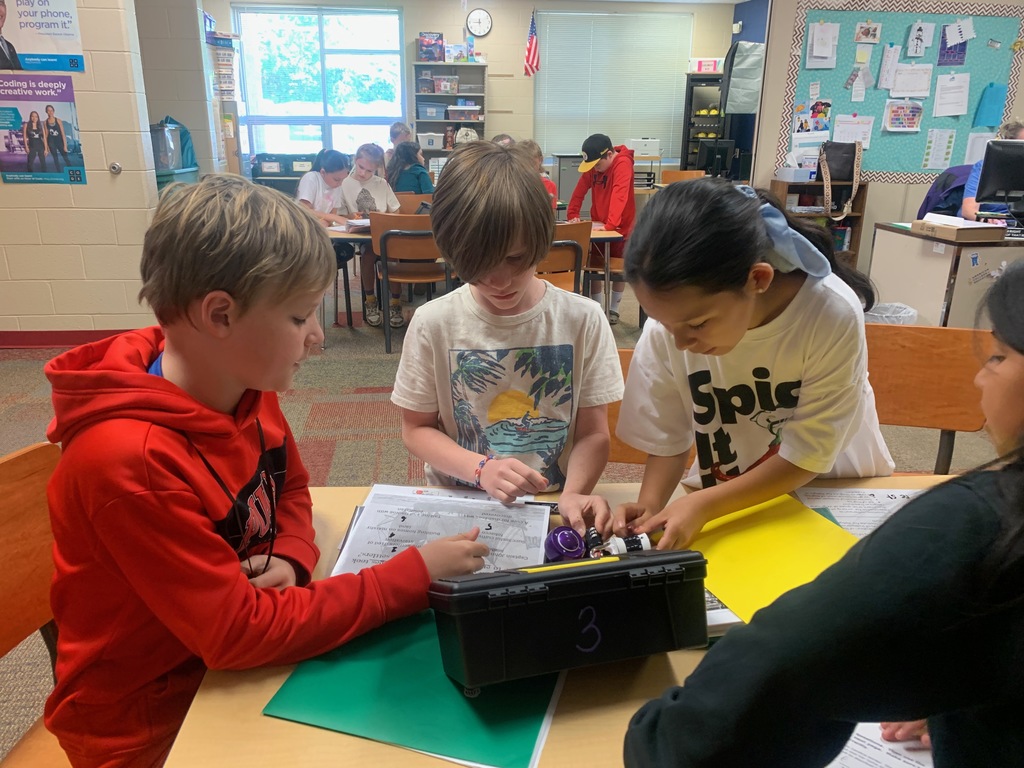 A small group of students gathers around a table, reading clues and manipulating locks on a black box as part of a classroom breakout activity. Additional students work in groups at tables behind them.