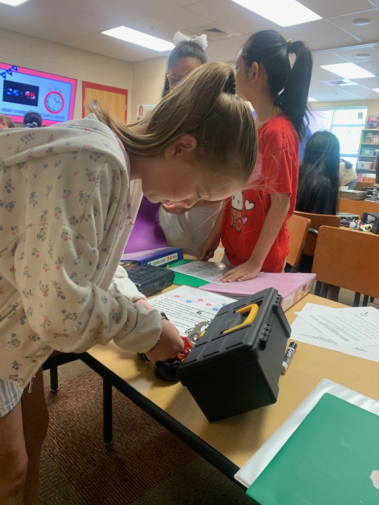 Students work together at a table in a classroom, focusing on a locked box and printed clues. One student leans in closely while using a combination lock as others observe and assist.