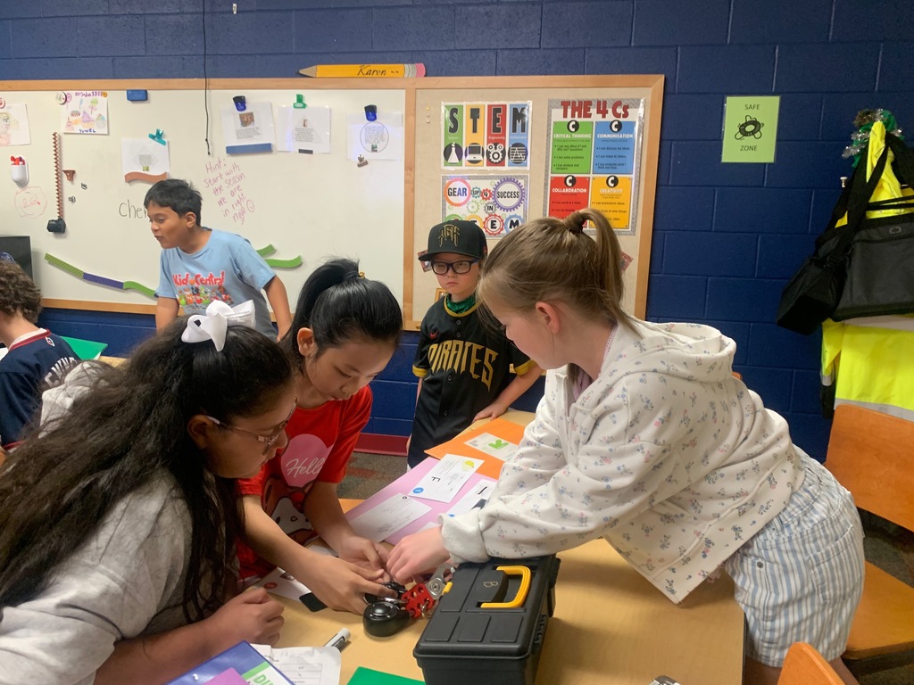 Four students stand around a table, working together to solve a lock puzzle using printed clues and materials. A classroom wall behind them displays STEM and “4 Cs” posters emphasizing critical thinking and collaboration.