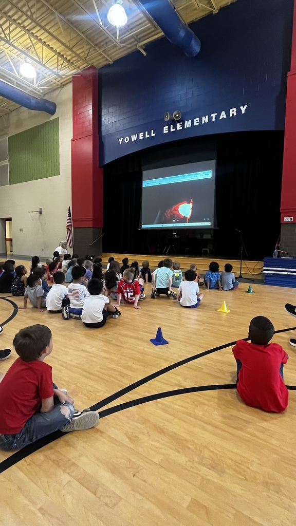 Students sit on the gym floor watching a live demonstration of egg candling projected onto a large screen on stage.