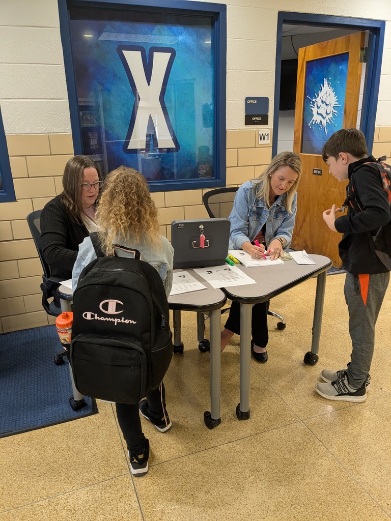 Two staff members sit at a table in a school hallway, checking forms and writing with highlighters while two students stand nearby with backpacks, participating in the “Tat-A-Teacher” activity outside the office.