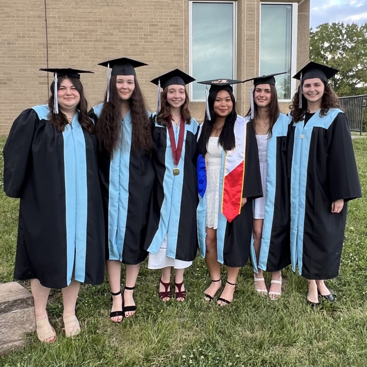 a group of six young women dressed in cap and gown standing on the grass in front of a brown, brick building.