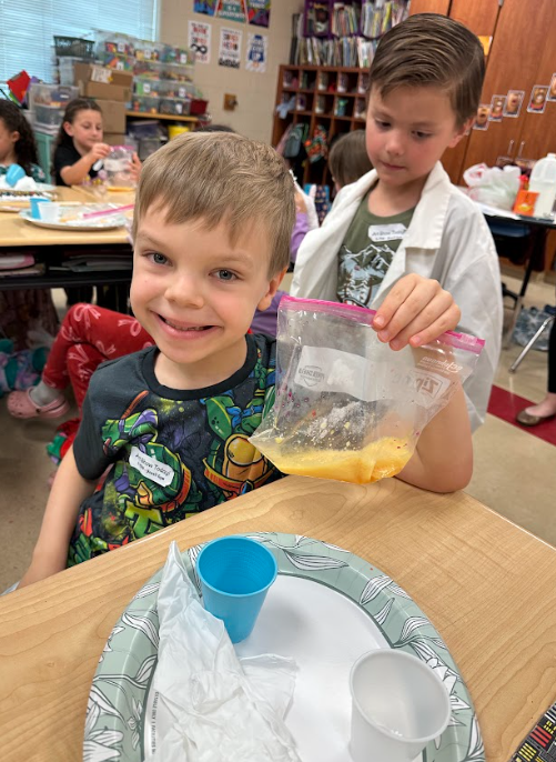 Young boy smiling at a classroom desk while another child holds up a plastic bag with a yellow liquid mixture during a hands-on science activity.