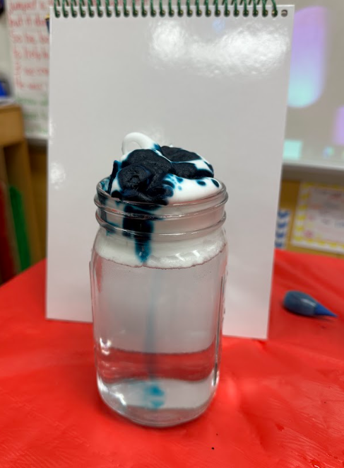 Glass jar filled with clear liquid topped with white foam and dark blue dye dripping downward, demonstrating a science experiment.