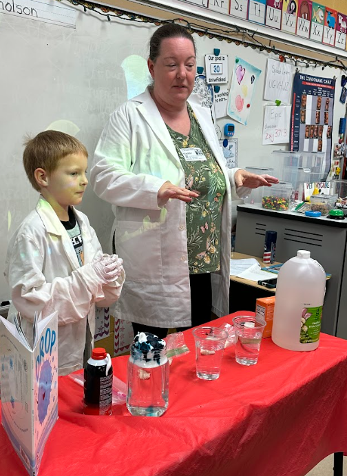 Teacher and student wearing lab coats stand behind a table with jars, cups, and liquids, preparing to demonstrate a science activity.