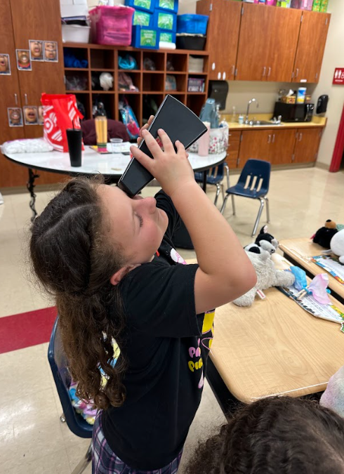 Student tilts a black container to their eye as if looking through it during a classroom experiment, with desks and supplies behind them.
