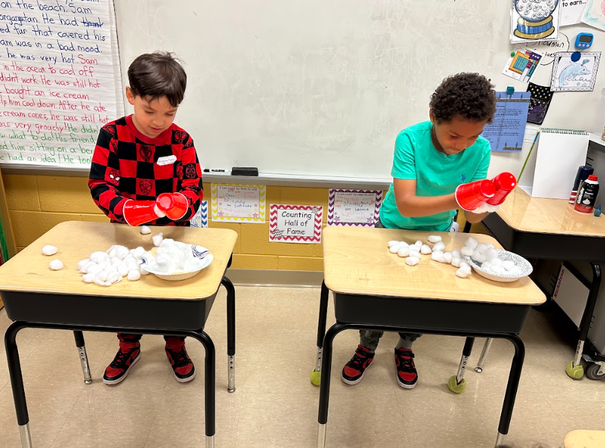 Two students at separate desks use red cups to move or stack white cotton balls as part of a classroom activity.