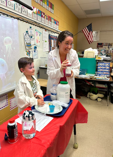 Teacher in a lab coat smiles while holding a bottle of liquid next to a student, both standing behind a table set up for a science demonstration.