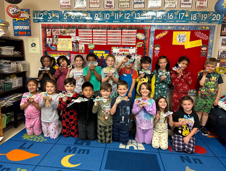 Large group of students pose together in a classroom holding small craft projects, smiling for a group photo in front of a decorated bulletin board.