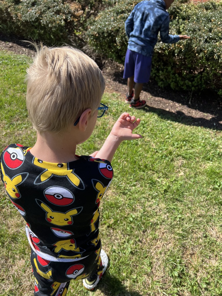 A young child wearing Pikachu-themed clothing holds a small green object in their hand while standing on grass, with another child nearby.